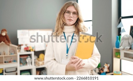 Young blonde woman preschool teacher holding books with relaxed expression at kindergarten