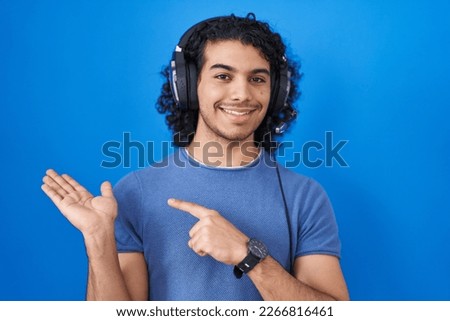 Hispanic man with curly hair listening to music using headphones amazed and smiling to the camera while presenting with hand and pointing with finger. 