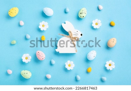 Sweet Colorful Easter Eggs, White Easter Bunny, daisy flowers on pastel blue background. Happy Easter greeting card concept. Flat lay, top view, copy space.