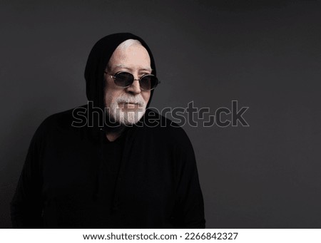 Charismatic elderly gray-haired man in sports black suit, hood and sunglasses, studio, gray background, place for text