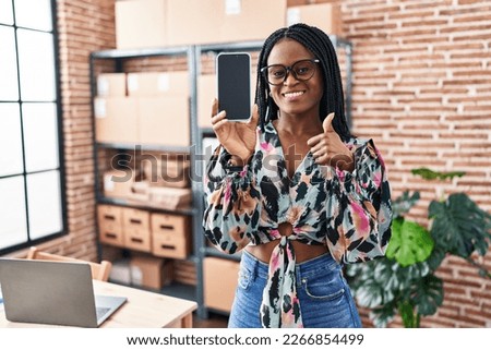 African woman with braids working at small business ecommerce showing smartphone screen smiling happy and positive, thumb up doing excellent and approval sign 