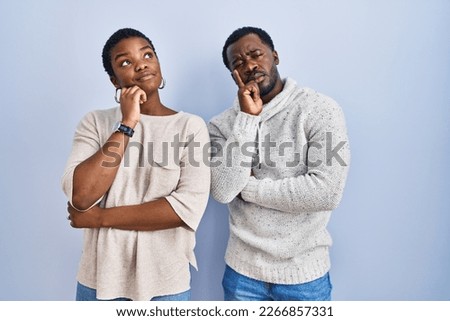 Young african american couple standing over blue background together with hand on chin thinking about question, pensive expression. smiling with thoughtful face. doubt concept. 