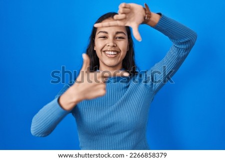 Young brazilian woman standing over blue isolated background smiling making frame with hands and fingers with happy face. creativity and photography concept. 