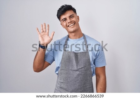 Hispanic young man wearing apron over white background waiving saying hello happy and smiling, friendly welcome gesture 