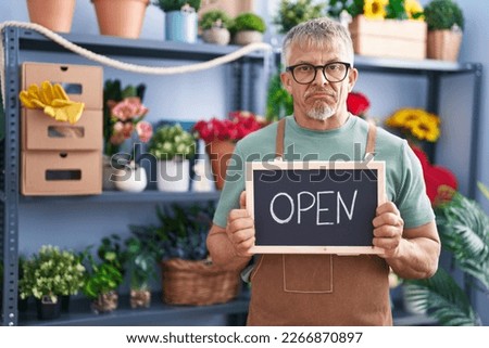 Hispanic man with grey hair working at florist holding open sign relaxed with serious expression on face. simple and natural looking at the camera. 