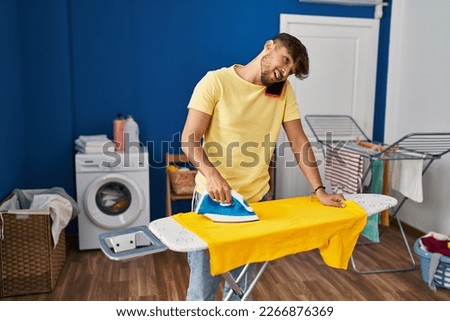 Young arab man talking on smartphone ironing clothes at laundry room