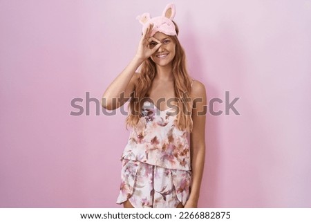 Young caucasian woman wearing sleep mask and pajama doing ok gesture with hand smiling, eye looking through fingers with happy face. 