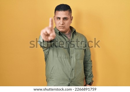 Hispanic young man standing over yellow background pointing with finger up and angry expression, showing no gesture 