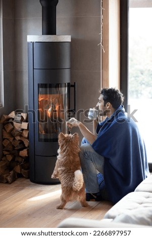 Young man sitting close to the fireplace with his cute joyful dog and warming in cold winter day
