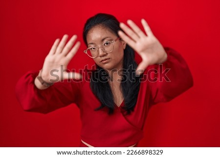 Asian young woman standing over red background doing frame using hands palms and fingers, camera perspective 
