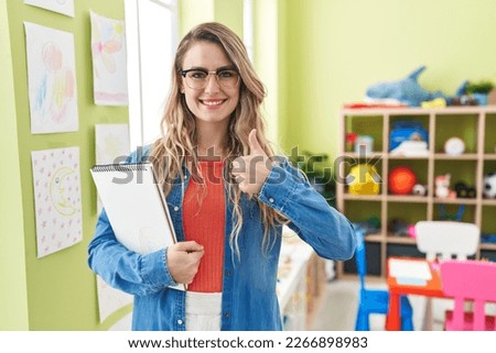 Young caucasian woman working as teacher at kindergarten smiling happy and positive, thumb up doing excellent and approval sign  Royalty-Free Stock Photo #2266898983