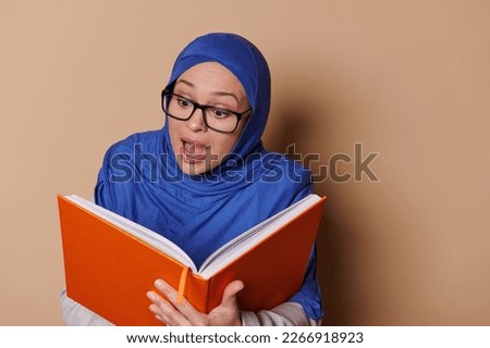 Amazed Arab Muslim woman wearing eyeglasses and blue hijab, expressing surprise and happy emotion while reading fairy tail, isolated on beige background. People. Education. Erudition. Learning concept
