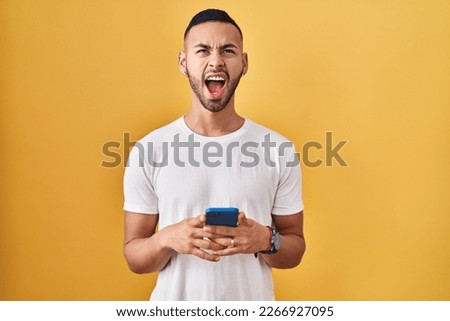 Young hispanic man using smartphone typing message angry and mad screaming frustrated and furious, shouting with anger looking up.  Royalty-Free Stock Photo #2266927095