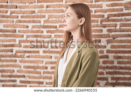 Young caucasian woman standing over bricks wall background looking to side, relax profile pose with natural face with confident smile. 