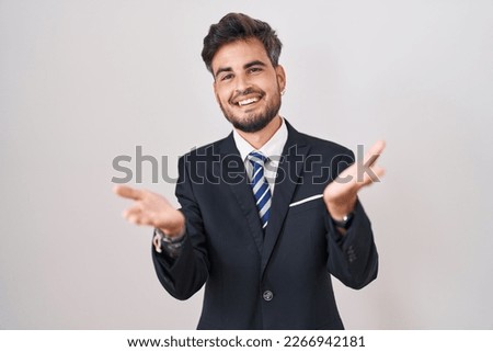 Young hispanic man with tattoos wearing business suit and tie smiling cheerful offering hands giving assistance and acceptance.  Royalty-Free Stock Photo #2266942181