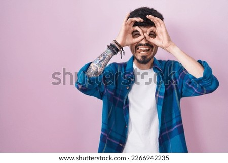 Young hispanic man with beard standing over pink background doing ok gesture like binoculars sticking tongue out, eyes looking through fingers. crazy expression. 