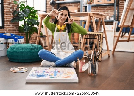 Hispanic woman sitting at art studio painting on canvas smiling making frame with hands and fingers with happy face. creativity and photography concept. 
