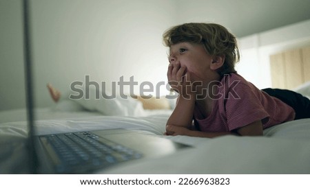 Young boy starring at media entertainment on laptop screen laying in bed at night. Hypnotized kid staring at blue glowing screen in the evening