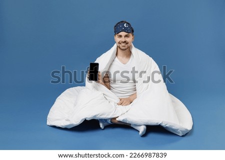 Full length young man in pajamas jam sleep mask rest relax home sit wrap under cover blanket duvet use mobile cell phone workspace blank screen isolated on dark blue background Night bedtime concept