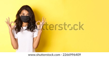 Concept of coronavirus, pandemic and lifestyle. Portrait of confident african-american girl, wearing black face mask to protect herself from covid-19, showing okay signs, yellow background.