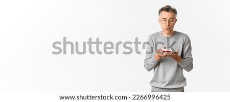 Portrait of attractive middle-aged man in grey sweater and glasses, celebrating his birthday, blowing a candle on b-day cake and making wish, standing over white background.