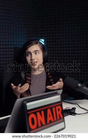 Young female with headphones and long braids clasping hands and looking away while sitting at table during podcast in recording studio