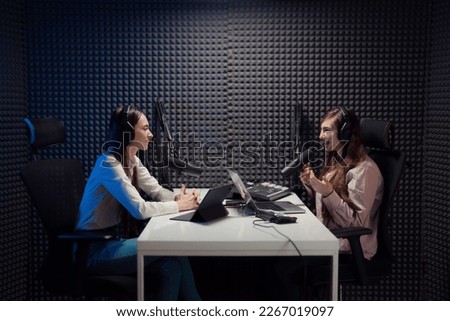 Cheerful female host and guest sitting at table with gadgets and and speaking with each other while recording podcast in modern studio