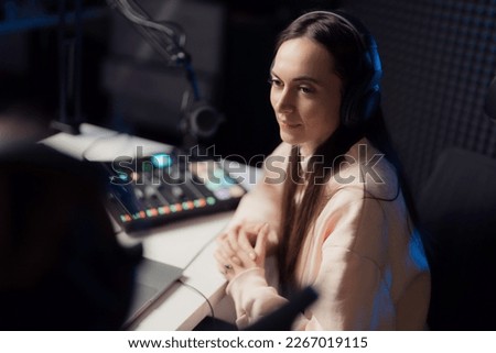 Young female with headphones clasping hands and looking away while sitting at table during podcast in recording studio