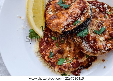 This close-up photo showcases a round serving of eggplant parmesan in a white flat bowl, complete with a lemon wedge. The textures and colors perfect for food or culinary design projects Royalty-Free Stock Photo #2267030815