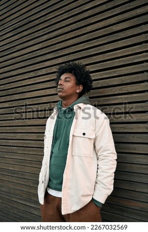 Cool young hipster gen z African American teen standing at wooden wall. Sensitive vulnerable ethnic rebel teenage boy breathing air with eyes closed feeling free or sad in city outdoors. Vertical