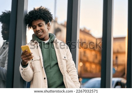 Cool smiling young African American teen guy holding mobile phone standing at glass city wall. Happy stylish hipster teenager tech user using cell outdoors, looking at cellular checking smartphone.