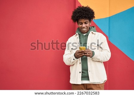 Happy young African American guy standing at color bright red wall outdoors using cell phone, looking at camera holding cellphone enjoying doing online ecommerce shopping in mobile apps, playing game.