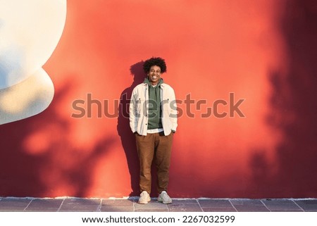 Happy African American teen guy having fun laughing on red wall lit with sunlight. Smiling cool ethnic gen z teenager model standing looking at camera, authentic full length shot outdoors.
