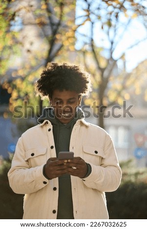Cool smiling young African American guy holding mobile phone standing with smartphone at big city sunny street. Hipster gen z teen boy using cellphone gagdet outdoors, looking at smartphone. Vertical