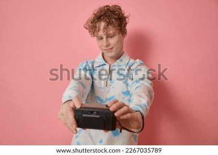 Horizontal studio shot of a young curly haired cute cheerful smiling boy in bright clothes taking selfie on film camera photo captured waist-up to remember his childhood against pink background.