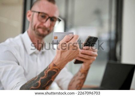 selective focus image, Thoughtful Caucasian man with glasses and tattoo on his arms holding a credit card and his smartphone at his desk. 