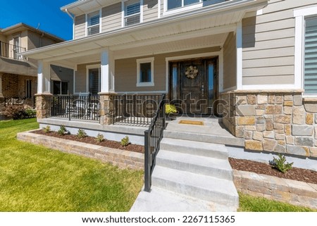 House facade with railings on the porch with wide dark wood front door with flower wreath. Two-storey house with wood and stone veneer siding beside the house with bricks on the left. Royalty-Free Stock Photo #2267115365