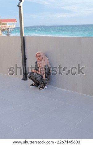 Portrait of young confident Asian woman sitting squatting smiling near beach wall