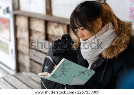 Asian woman traveler writing journey memories in diary book during travel small town in snow day. Attractive girl resting at wooden seat pavilion covered in snow during travel Japan on winter vacation