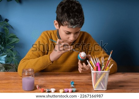 Creative 10-year-old caucasian boy decorating a white Easter egg at his desk with watercolor paint and brushes, surrounded by more Easter eggs and art supplies. Great for Easter-themed designs