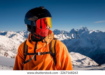 Portrait of a young man in a ski mask, stands in a ski resort against the backdrop of mountains and blue sky
