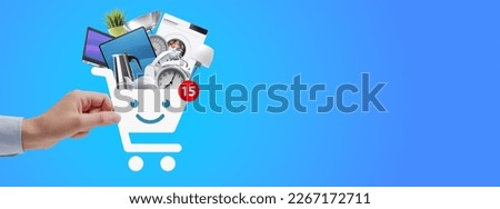 Woman holding a smiling shopping cart icon filled with household goods and electronics, online shopping concept Royalty-Free Stock Photo #2267172711