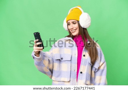Young woman wearing winter muffs over isolated chroma key background making a selfie