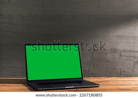 Chroma key green screen, angled view laptop on wooden table in front of concrete wall. Table top shot of interior space with window light effect.