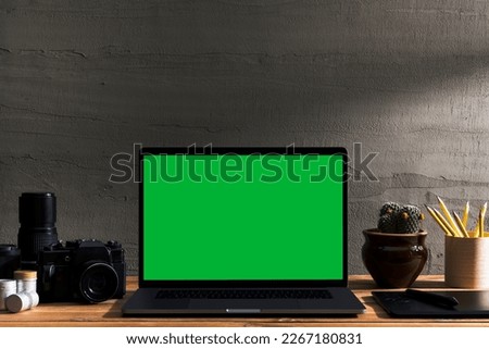 Chroma key green screen laptop with photography equipment on wooden table in front of concrete wall. Table top shot of interior space with window light effect.