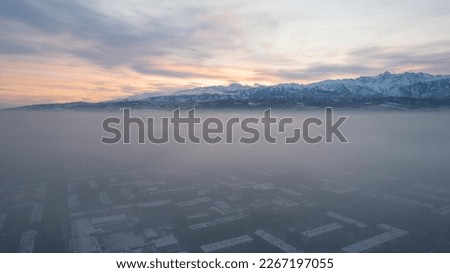 Dawn over the city in fog and smog. Mountain view. A light haze hangs over the city, cars drive, signs glow. Clouds, sun rays and snowy mountains are visible in the distance. Almaty, Kazakhstan