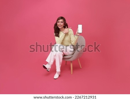 Happy young Asian woman showing empty screen mobile phone. While her sitting on chair isolated on pink studio background. Royalty-Free Stock Photo #2267212931