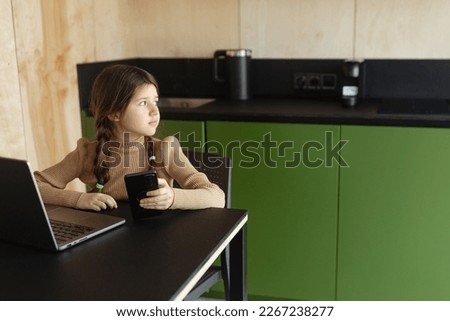 A girl at online home education sits at a laptop and a phone on a green background, inside, caucasian, ukrainian child