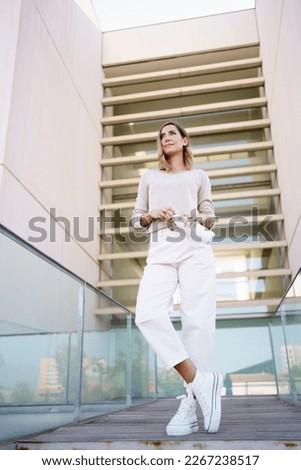 Low angle of woman in stylish outfit opening zero waste bottle of water and looking away while standing on path outside contemporary building Royalty-Free Stock Photo #2267238517