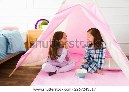 Happy little girls in pajamas playing during a fun sleepover eating popcorn while talking in a pink teepee Royalty-Free Stock Photo #2267243517